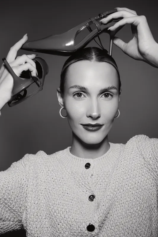 Caucasian woman with fair skin and dark hair holds high heel shoes near her head, wearing knitted sweater and earrings.