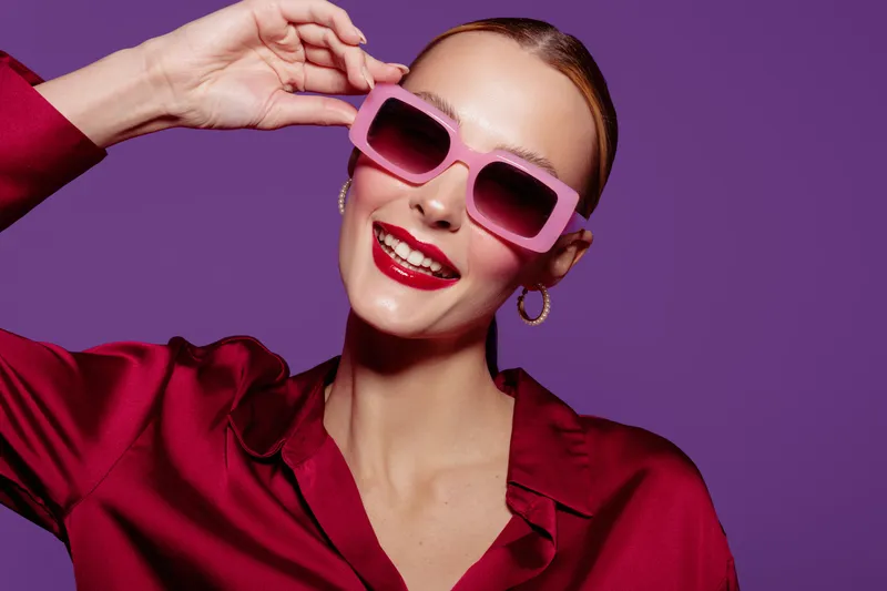 Smiling woman in red blouse wearing pink sunglasses poses against purple background. Fashion portrait with bright makeup and joyful expression. Concept of style, beauty, and confidence.