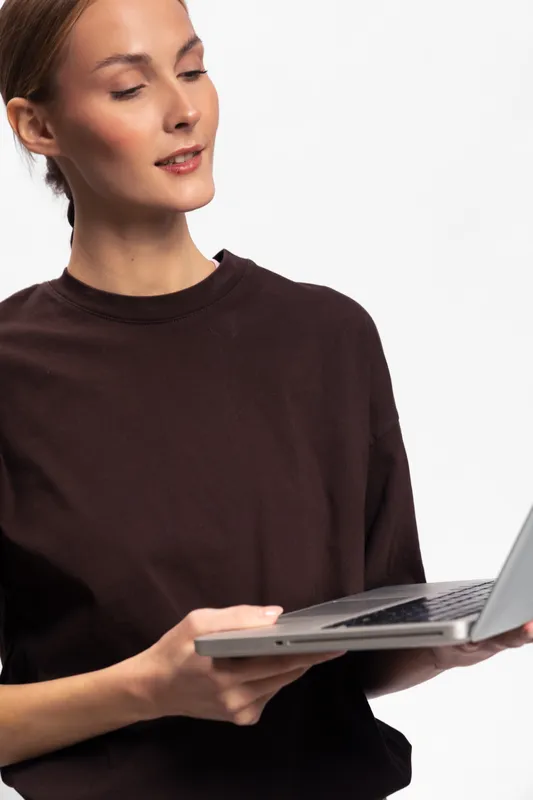 Young woman in dark sweater holds laptop and looks down with slight smile. Isolated on white background. Concept of technology, work, and casual lifestyle.
