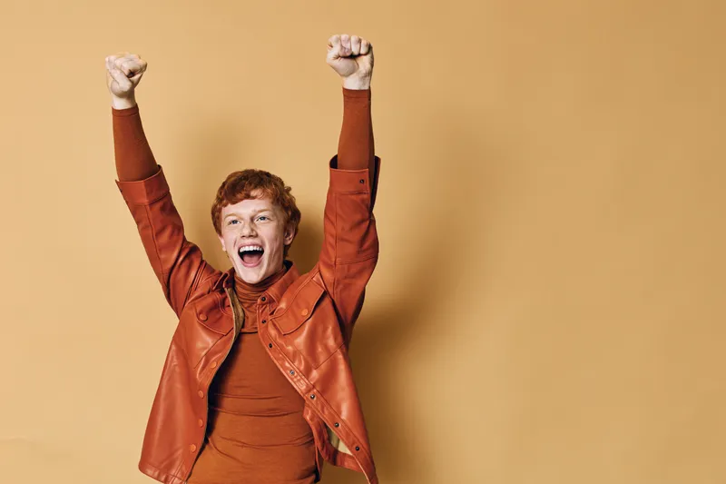 Young man with red hair wearing brown jacket and sweater celebrating with raised hands and open mouth on beige background for joy and success expression.