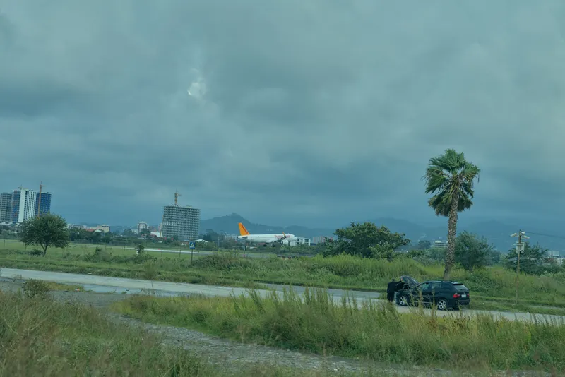 Airfield scene with airplane in distance, wet runway, lone car on grassy verge, palm tree, tropical landscape under stormy clouds, moody atmosphere, suggesting travel, transit, or transportation