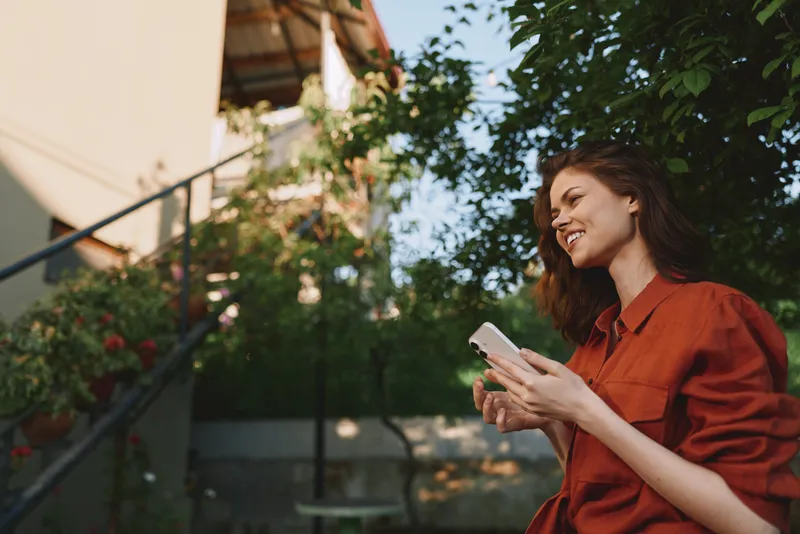 Woman lifestyle portrait outdoors, smiling while using a phone, wearing a red shirt in a garden setting, relaxed cheerful mood, natural light, candid moment
