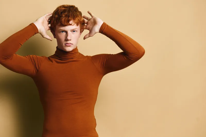 Young man with red hair wearing brown turtleneck sweater posing with hands near head against plain beige background for fashion or portrait concept.