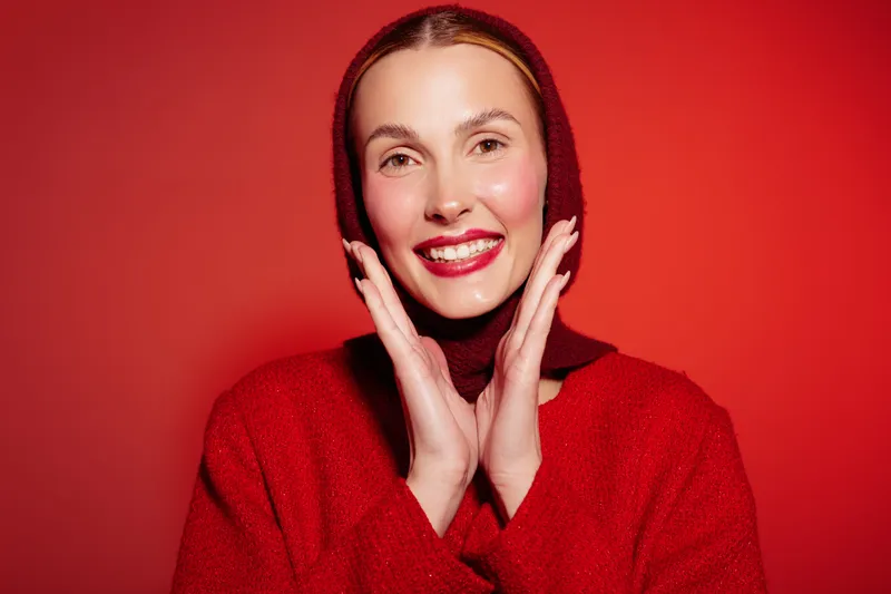 Smiling woman in red sweater and matching headscarf poses with hands near face against red background. Concept of fashion, winter style, beauty, and cheerful mood in studio lighting.