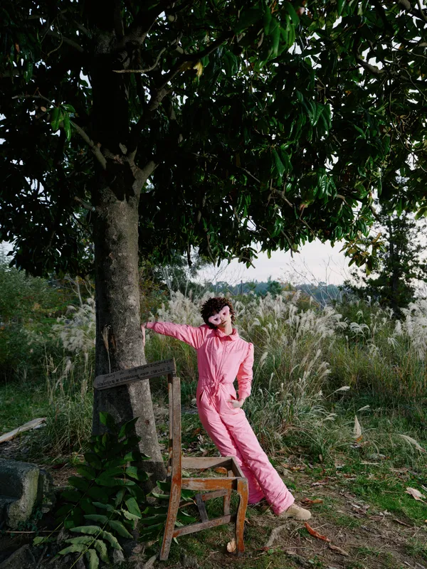 Outdoor woman in a pink jumpsuit leans against a tree in a rustic garden, a relaxed pose under leafy shade with natural light and calm surroundings, conveying playful style and outdoor serenity.
