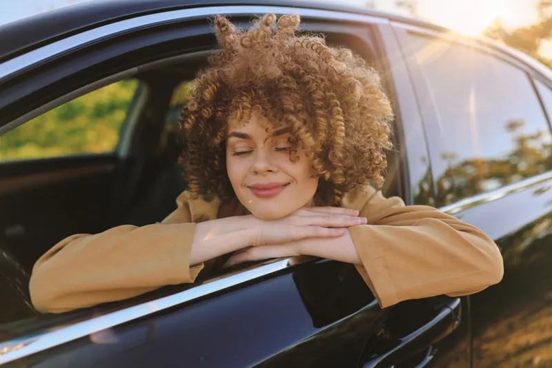 Smiling woman with curly hair resting her arms on a car window, bathed in warm sunset tones. Outdoor lifestyle scene, relaxed mood, retro film color, natural lighting, peaceful moment.