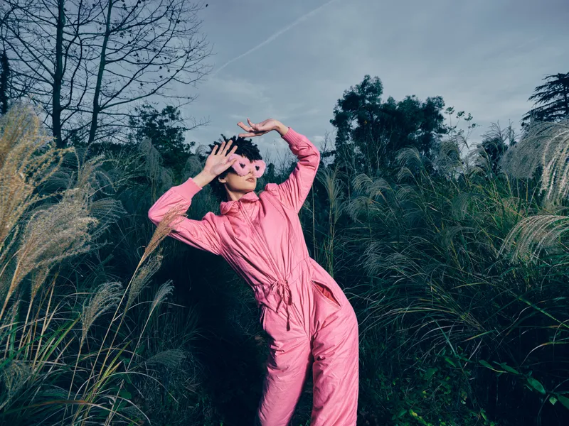 Outdoor fashion scene with a person in a pink jumpsuit posing among tall grasses, sunglasses on, dynamic pose, vibrant mood, nature backdrop and energetic lifestyle vibe.