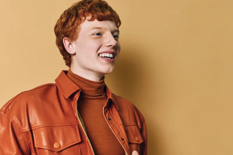 Young man with red hair and freckles smiling wearing brown leather jacket and turtleneck standing against beige background in studio portrait.