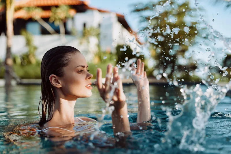 A woman in a pool splashes water, concentrating as droplets fly around under bright outdoor sunlight, capturing a moment of playful movement, refreshing recreation, and summer leisure in a vibrant