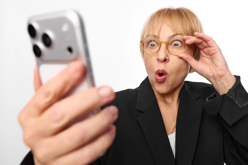 Woman selfie smartphone surprised glasses office portrait, surprised expression in blazer. White studio background and soft light show shock and digital online impact communication.