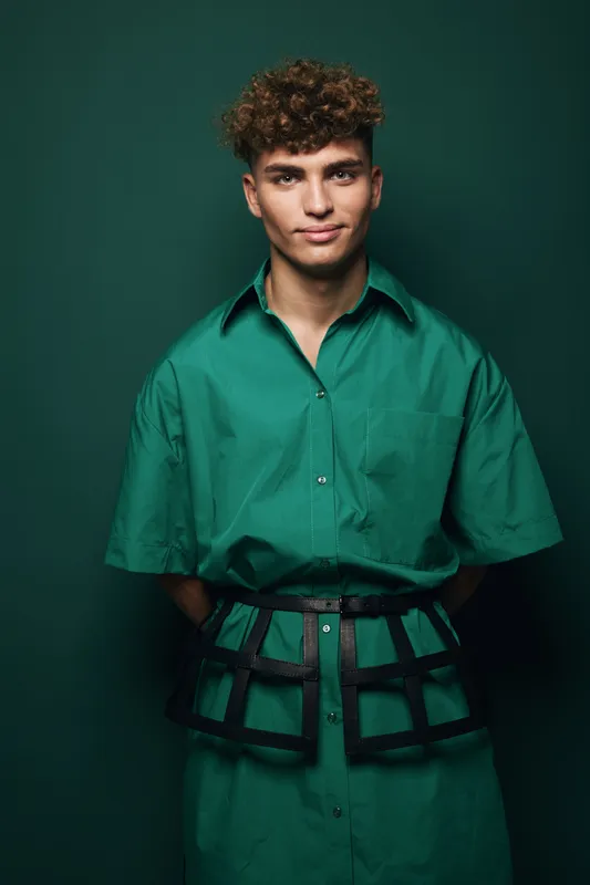 Young man with curly hair wearing green shirt dress and black harness belt posing against dark green background in studio. Fashion portrait with confident expression and casual style.