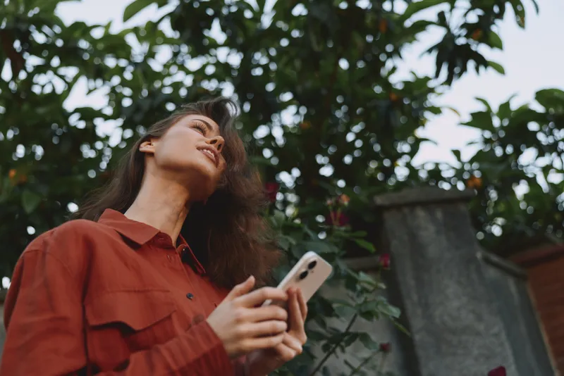A woman stands outdoors in a red shirt, looking up with a phone in hand. Natural light, candid mood, everyday lifestyle moment suggesting curiosity and connection