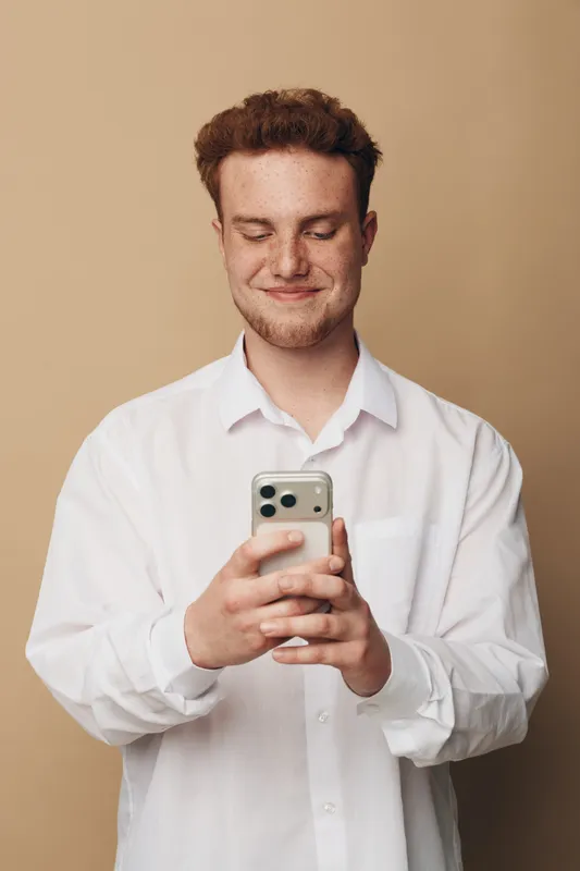 Smiling young man with curly hair wearing white shirt looks at smartphone screen, standing against beige background. Concept of technology, communication, and casual lifestyle.