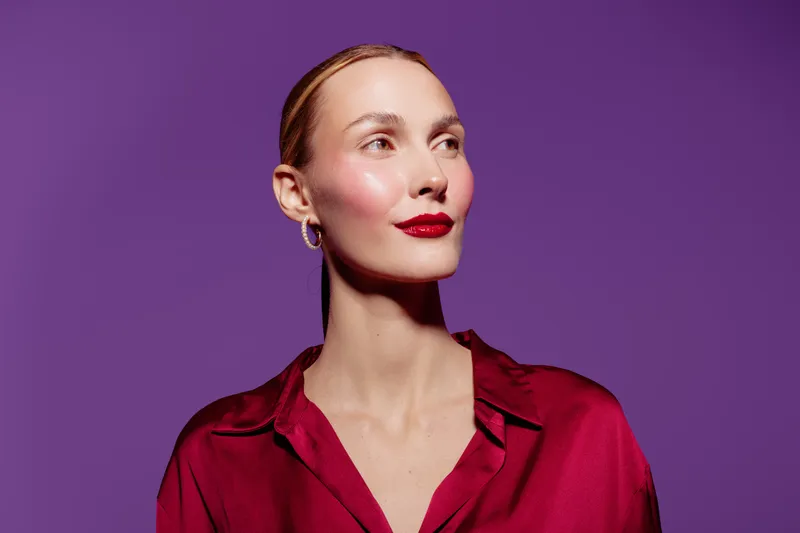 Confident woman with red lipstick and shiny skin looks aside on purple background. Studio portrait of elegant female model in red blouse with natural makeup and smooth hairstyle.