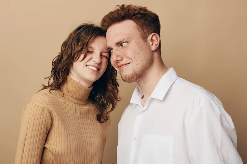 Smiling young couple with curly hair and red hair stand close together on beige background. They show affection and happiness in casual clothing. Concept of love and friendship.