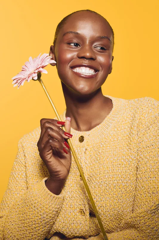 Smiling dark skinned African woman in yellow sweater holds pink flower on orange background. Concept for happiness, beauty, natural skin, and positive mood with floral element.