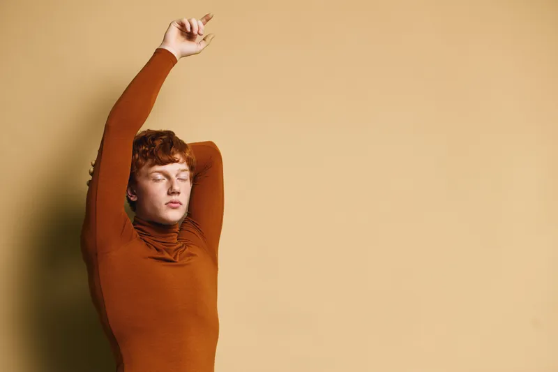 Woman with red hair stretching arms upwards with eyes closed against beige background. Relaxation, calm, casual clothing, turtleneck sweater, indoor portrait of young adult.
