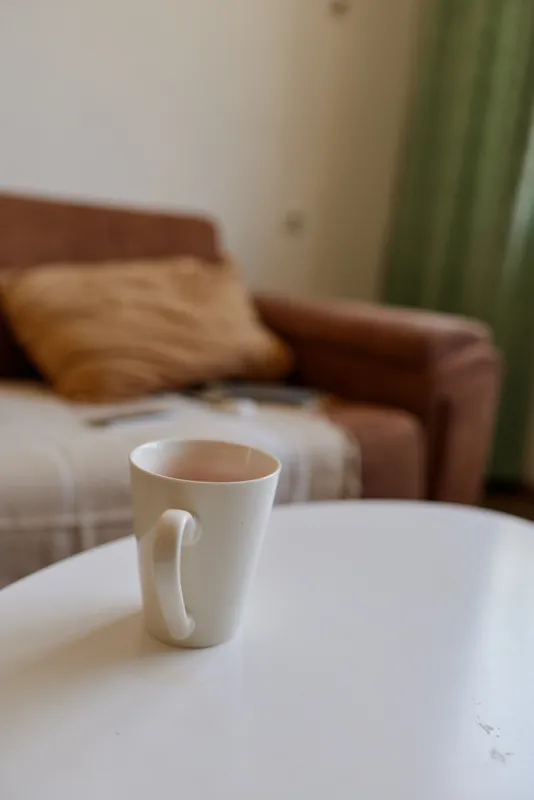 A white ceramic mug sits alone on a round white table in a cozy living room, inviting a quiet moment of reflection and simple daily calm.
