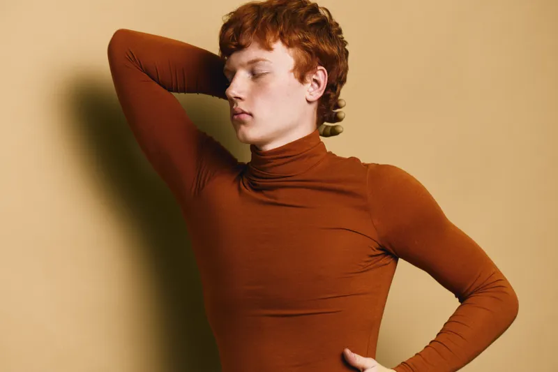 Young man with red hair in brown turtleneck posing with eyes closed and hand behind head against beige background for fashion portrait and style concept.