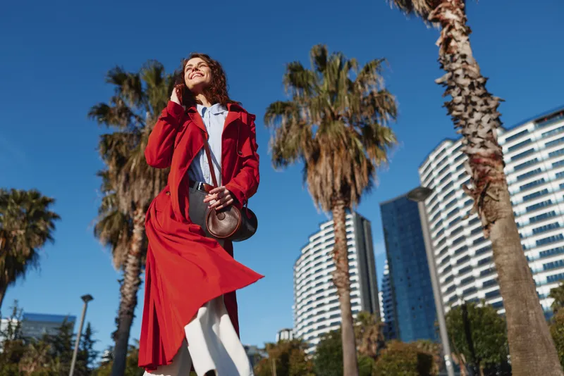 Young woman in a red coat walks confidently outdoors on a sunny day with palm trees and modern buildings in the background.