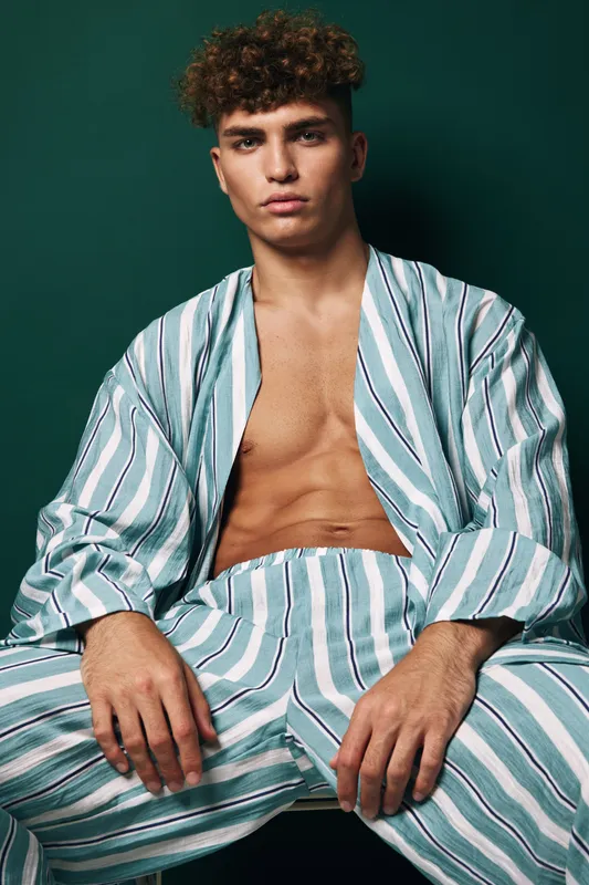 Young man with curly hair wearing striped open shirt and matching pants sitting against dark green background with serious expression and relaxed posture.