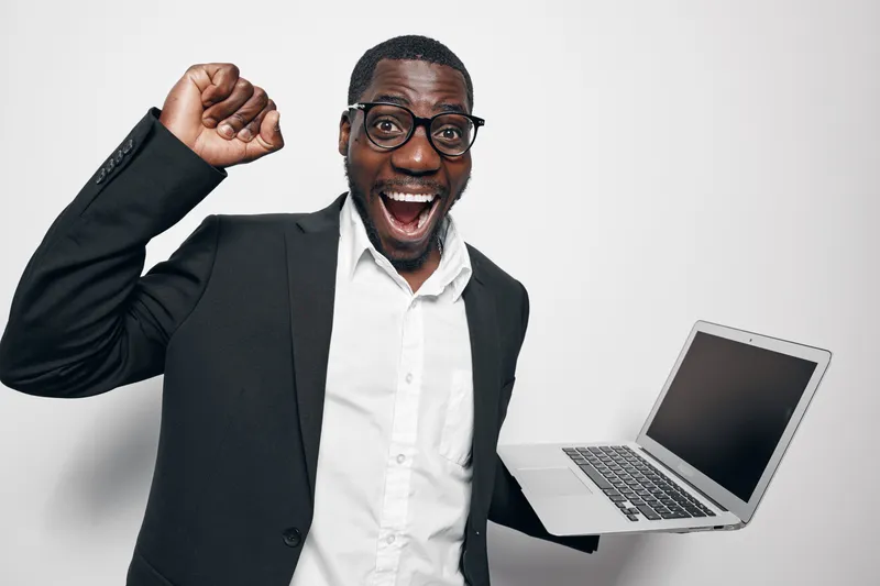 African American man with dark skin wearing glasses and black suit holding laptop celebrating success with raised fist and excited expression on white background in studio.