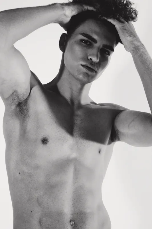 Young man with curly hair posing shirtless in black and white studio portrait, hands in hair, showing muscular torso and confident expression with soft lighting.