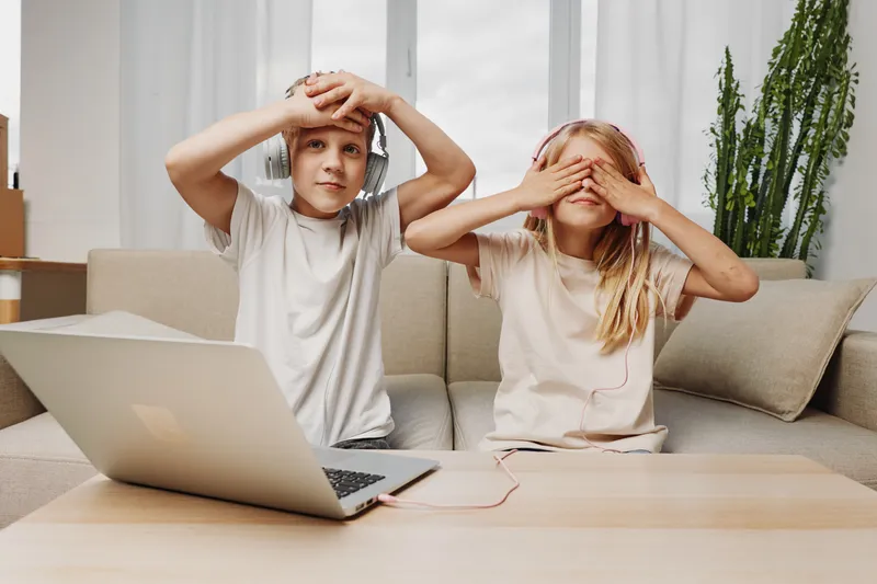 Two children sitting on a sofa with laptop, boy listening with headphones and girl covering eyes playing a game, casual home interior background.