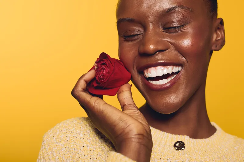 Close-up of smiling dark skinned African woman holding red rose near face on yellow background. Concept of beauty, happiness, natural skin, floral, and joyful emotions portrait.