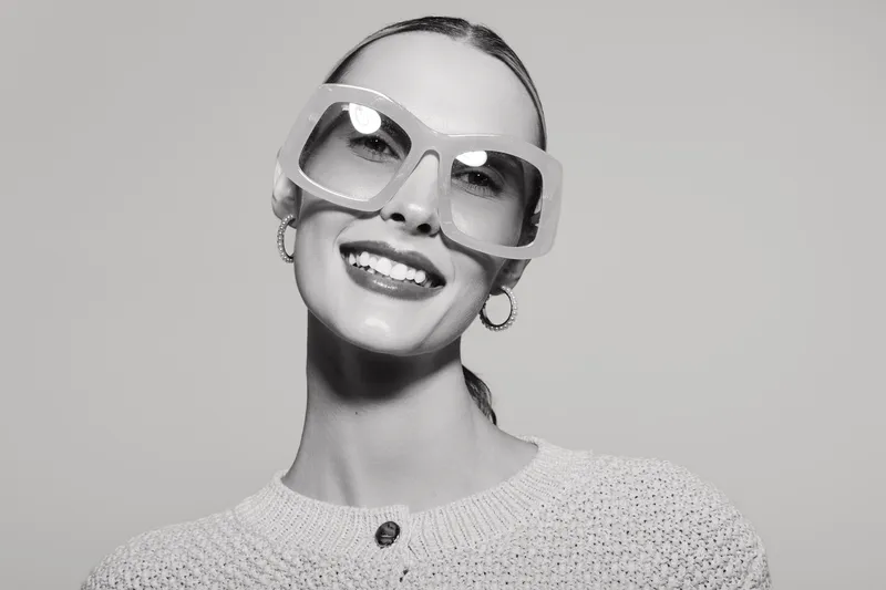 A smiling caucasian woman with fair skin wearing large glasses and a knitted sweater, isolated on white background.