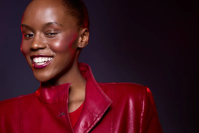 Close-up of happy African woman with dark skin wearing red leather jacket smiling confidently against dark background. Concept of fashion, beauty, and positive mood.