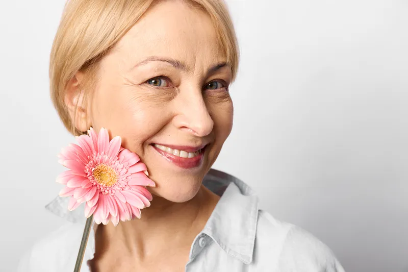 Woman portrait smiling blonde holding a pink gerbera flower near cheek, gentle smile, soft light, close up on white background, minimal composition, wellness mood, calm
