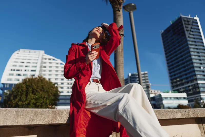 A woman in a red coat and white pants sits on a concrete ledge, looking up while holding a smartphone against a clear blue sky in a bright urban setting.