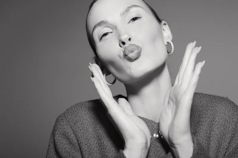 Young white woman with pale skin and earrings blows kiss with hands near face, isolated on grey background.