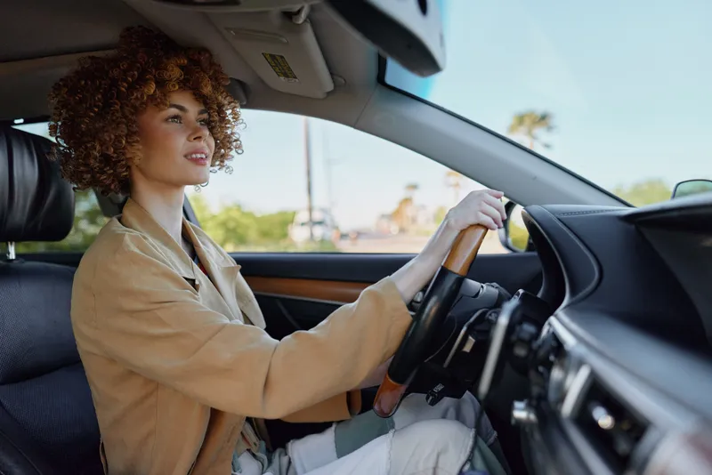 A woman driver with curly hair sits behind the wheel in a sunlit car, hands on the steering wheel, calm focus. Lifestyle portrait exploring independence, travel, and everyday commuting with retro