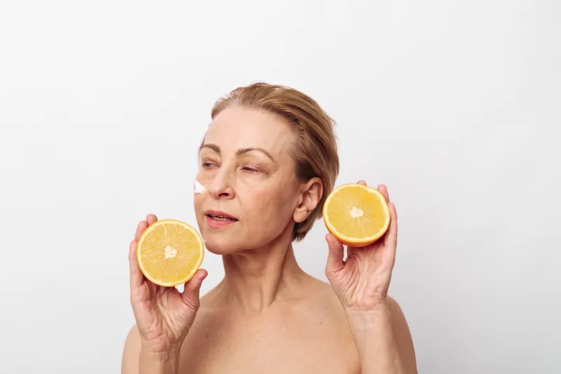 Mature woman with short hair holds two halves of fresh orange fruit near her face on white background. Concept of healthy lifestyle, natural beauty, and skincare with citrus.