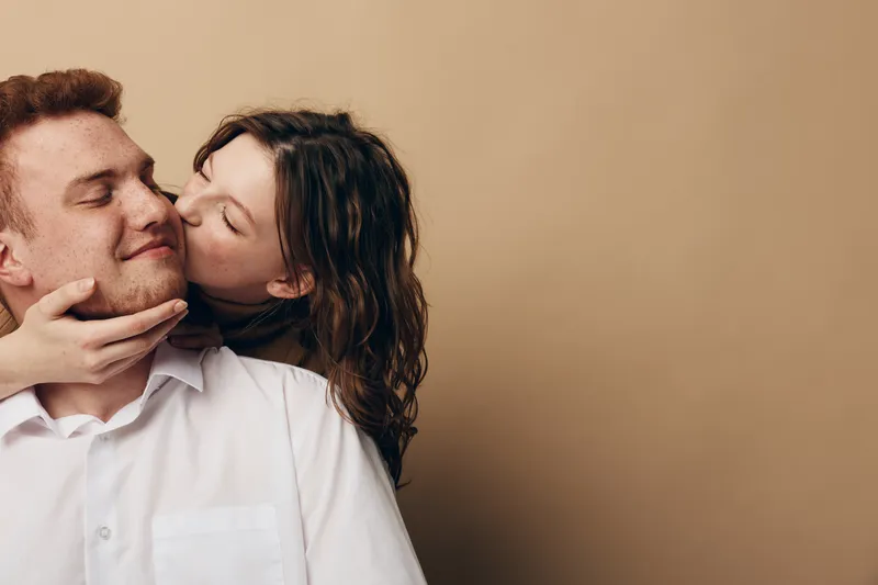 Young couple in casual clothes share a tender moment, woman kissing man on cheek with closed eyes. Warm beige background creates intimate and loving atmosphere, concept of affection and romance.