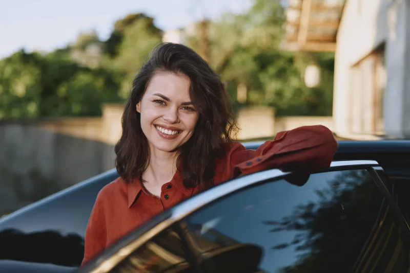 Woman lifestyle portrait smiling beside a car in daylight, casual red shirt, outdoor setting, relaxed confident mood, candid everyday moment of joy and freedom