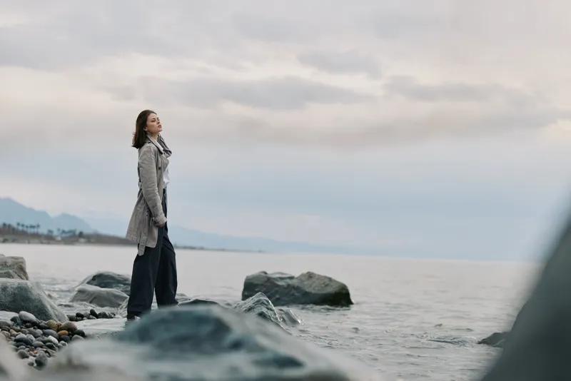 Young woman in a long coat stands on rocky shore looking at the sea under cloudy sky. Concept of contemplation, nature, and calm outdoor moment near water.