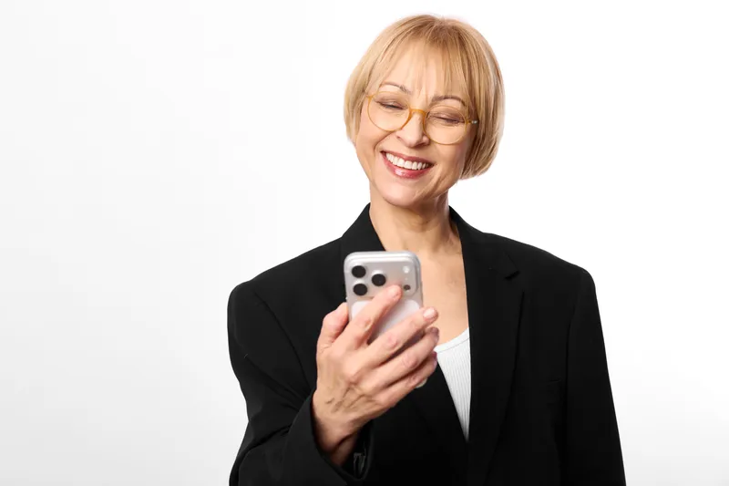Smiling elderly woman in black blazer holds a smartphone, portrait on white background, communication through technology, glasses visible, confident mobile lifestyle