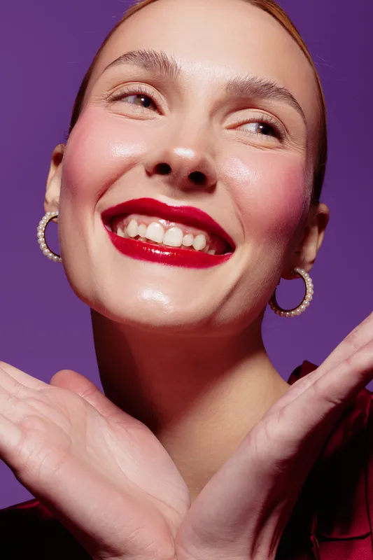 Smiling woman with red lipstick and hoop earrings poses with hands near face on purple background. Close-up portrait with natural skin glow and joyful expression. Concept of beauty and happiness.