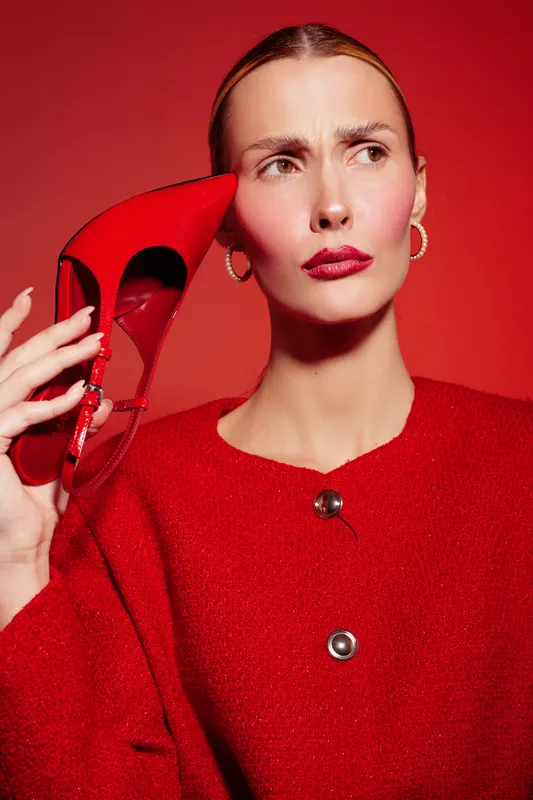 Stylish woman in red sweater holds red high heel shoe near face, looking confused. Red background and matching lipstick create bold fashion and beauty concept.