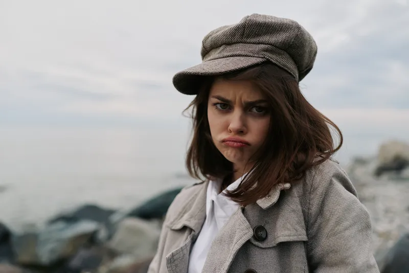 Young woman in a grey coat and cap shows a sad expression near rocky shore with cloudy sky. Concept of loneliness, disappointment, and emotional struggle outdoors in cold weather.