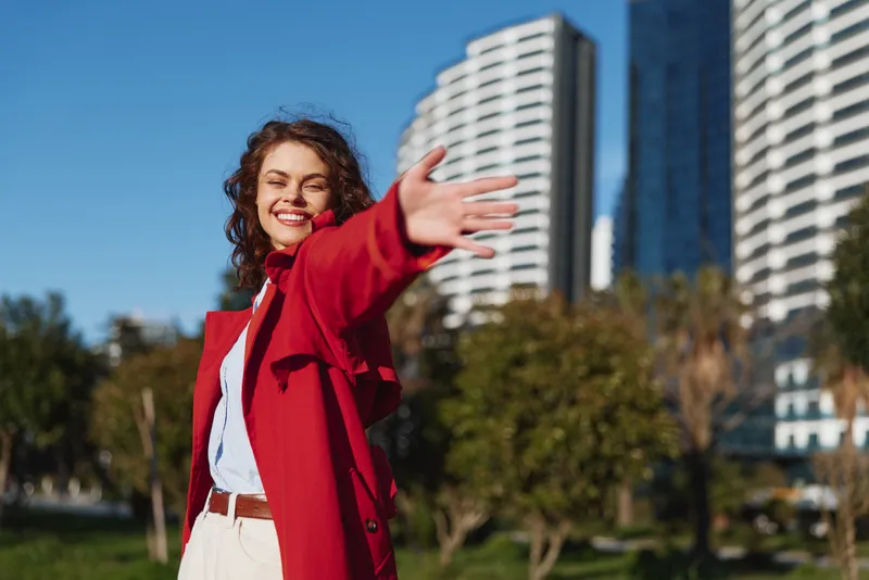 Young woman in a red coat smiles joyfully and reaches her hand toward the camera while standing in a sunny urban park with modern buildings in the background.