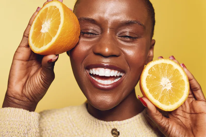 Happy African woman with dark skin holding two orange halves near her face smiles with closed eyes on yellow background. Concept of healthy lifestyle, freshness, and natural beauty.