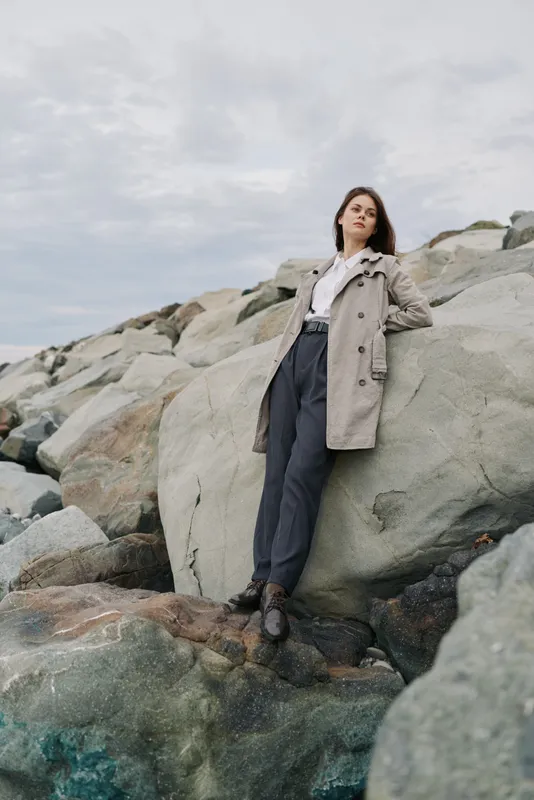 Young woman in trench coat leans on large rock by rocky shore under cloudy sky. Casual fashion and nature concept with relaxed pose and serene mood in outdoor setting.