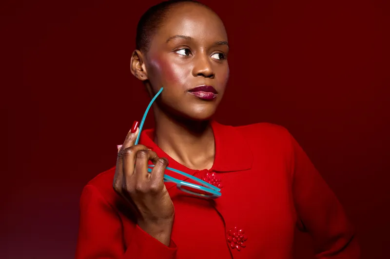 Confident black woman with dark skin and short hair wearing a red sweater holds blue glasses near face on red background. Concept of fashion, style, beauty, and modern portrait.
