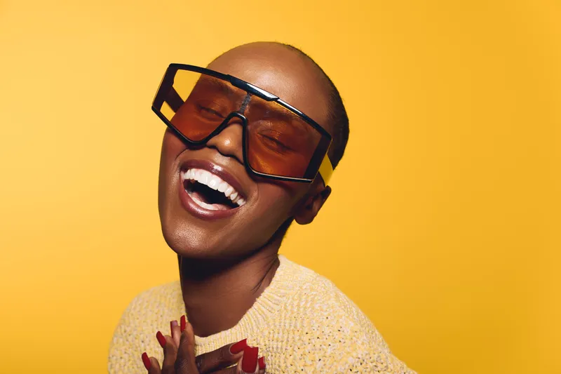 Happy dark skinned African woman with bald head wearing large sunglasses and beige sweater smiles with closed eyes on yellow background. Concept of joy, fashion and beauty.