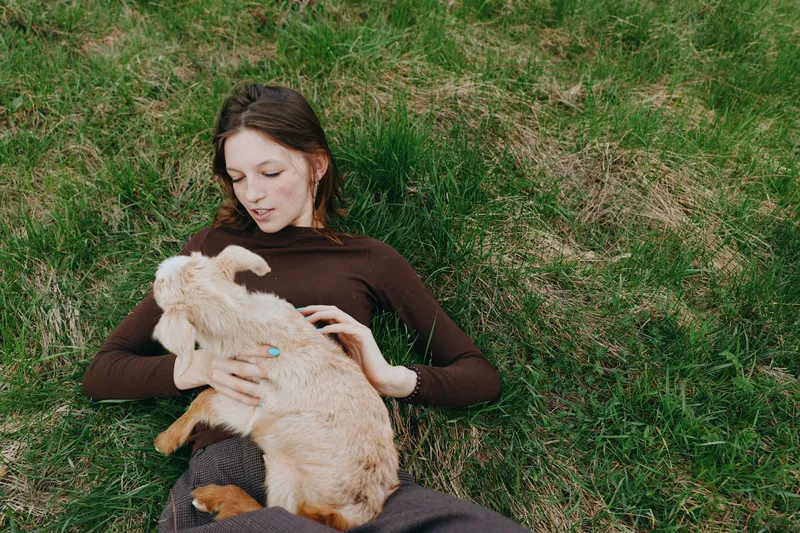 Young woman with long brown hair lies on grass holding a baby goat in her lap during a relaxed outdoor lifestyle moment in nature.