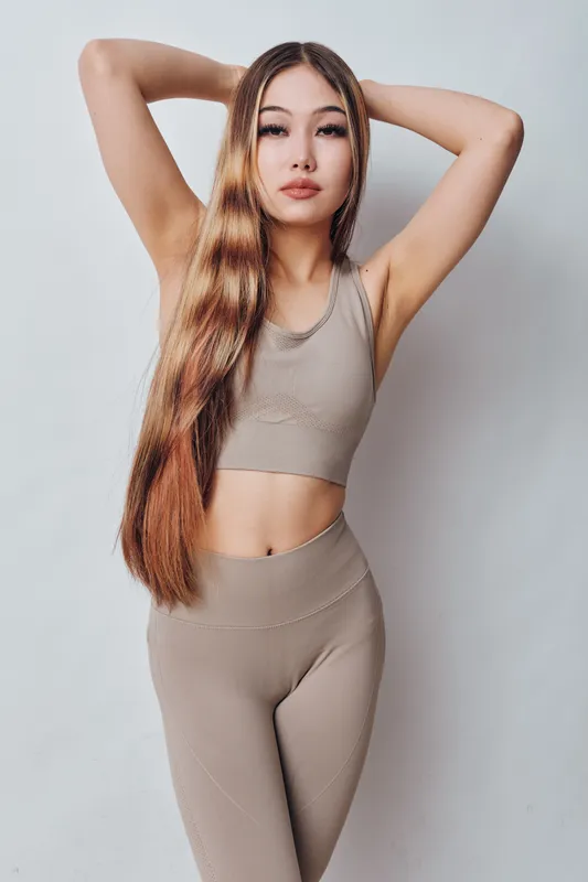 Young woman posing in beige fitness outfit with long hair and raised arms against white background. Casual activewear, confident model with natural makeup and healthy skin tone.
